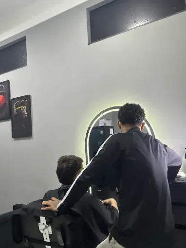 Dentro da barbearia, um barbeiro atendendo um cliente na cadeira, com espelho iluminado ao fundo